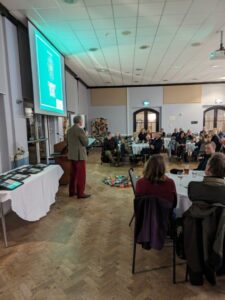 Michael Winter giving a talk at the Wild About Devon Awards 2023 with the certificates, audience, and colourful mossy carpet in view. Photo credits Kate Hind, DCC.